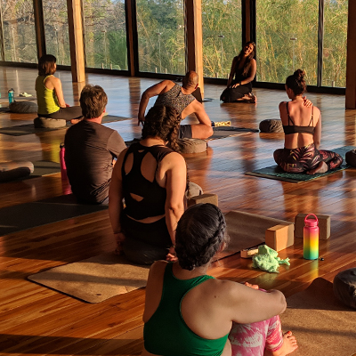 A yoga glass where adult students are sitted and listening to a female yoga teacher